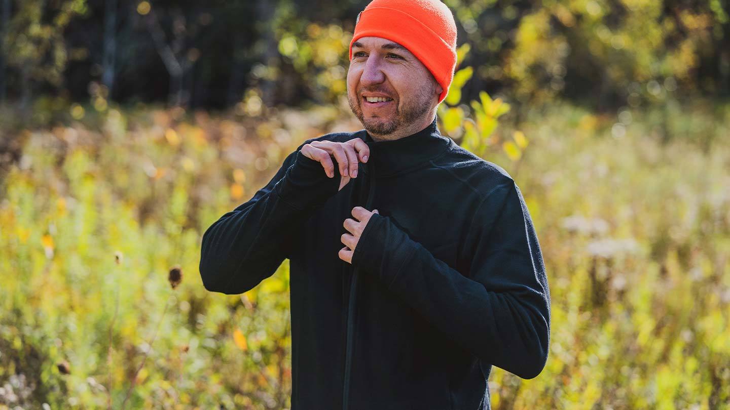 man in field putting on minus33 merino wool wilderness expedition full zip mid layer shirt and wearing beanie in blaze orange