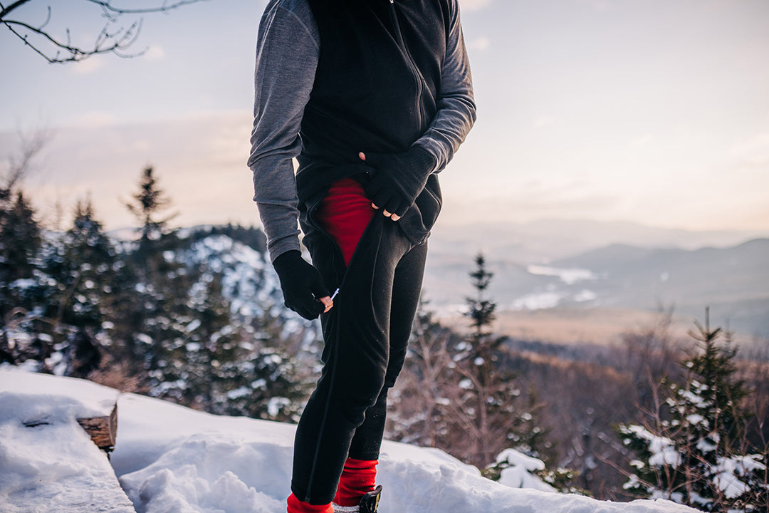 man ontop of a mountain zipping up his expedition weight zip off pants with midweight base layer bottoms underneath