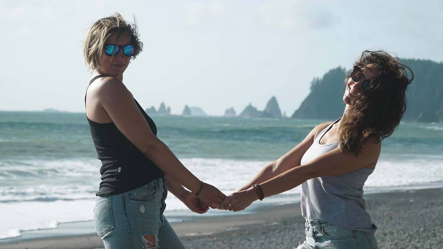 two women holding hands on the beach while wearing minus33 merino wool micro racerback tank tops