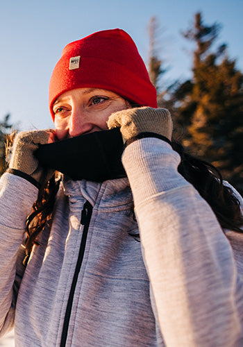 woman winter hiking pulling up her neck gaiter wearing a 100% merino wool beanie and merino wool fingerless gloves
