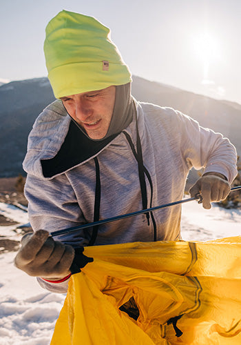 man setting up a tent on a mountain in the winter wearing a minus33 kodiak fleece hoodie and a midweight 100% merino wool neck gaiter with a high vis beanie