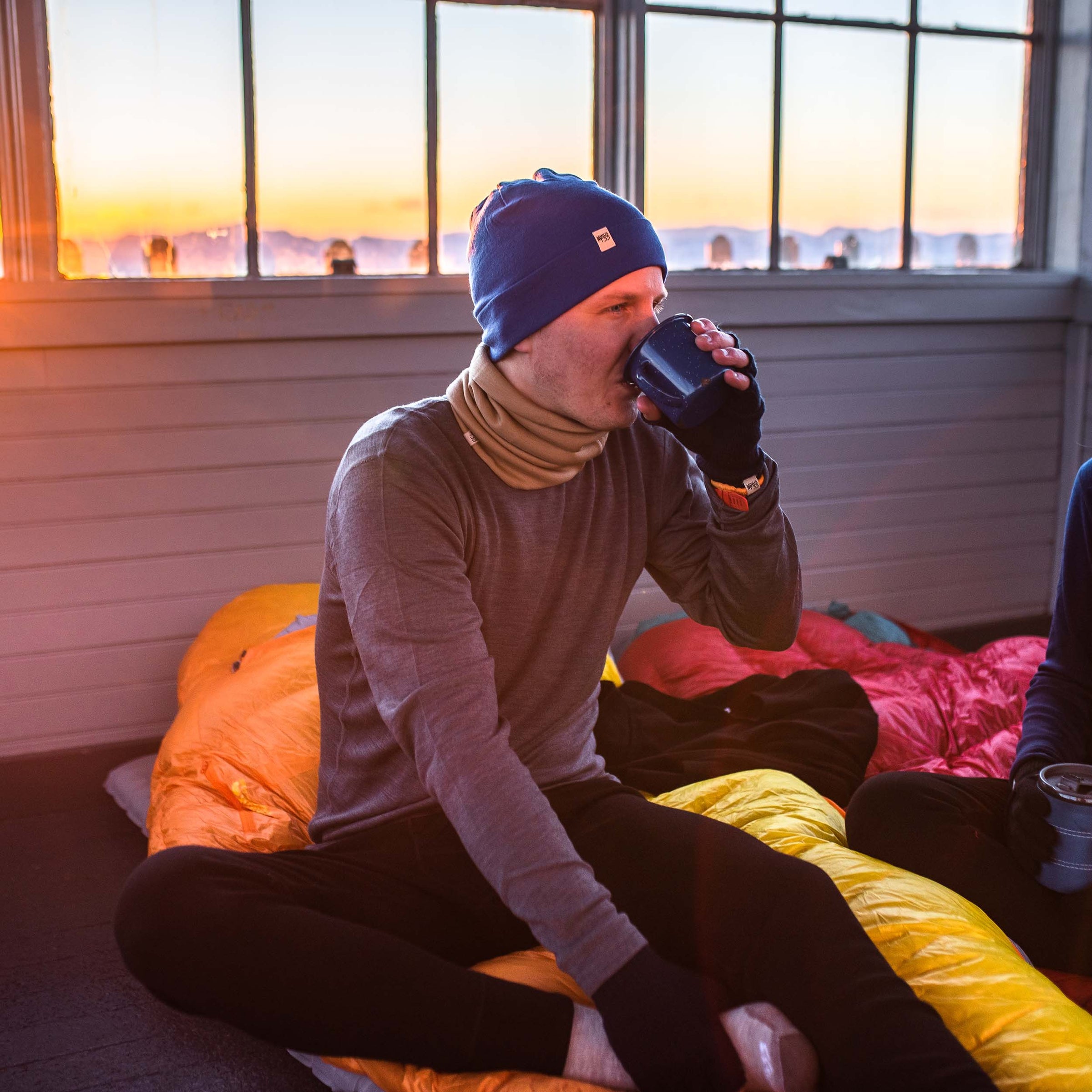 man drinking coffee while camping in the winter in a hut wearing midweight base layer best seller pair